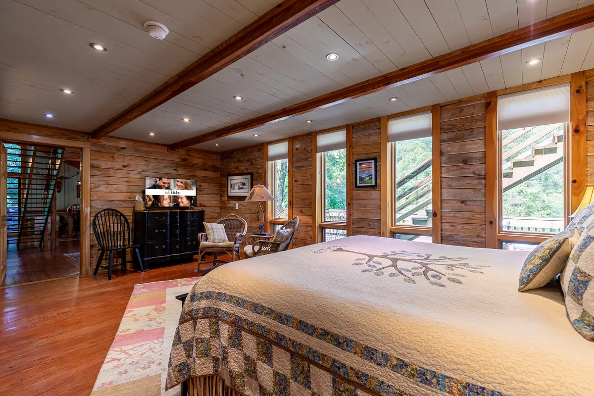 A bright, comfortable bedroom with a large bed adorned with a cozy quilt, natural wooden furniture, a television, and windows that flood the space with daylight and offer glimpses of nature outside.