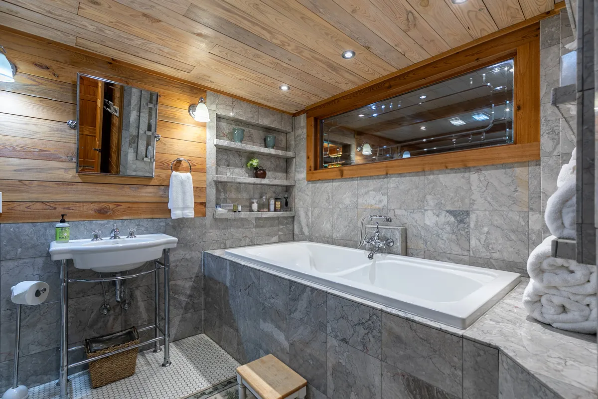 A luxurious bathroom with a freestanding soaking tub set against elegant marble walls, wooden shelves for storage, and a large mirror. The space feels spa-like and calming, ideal for relaxation and self-care routines.