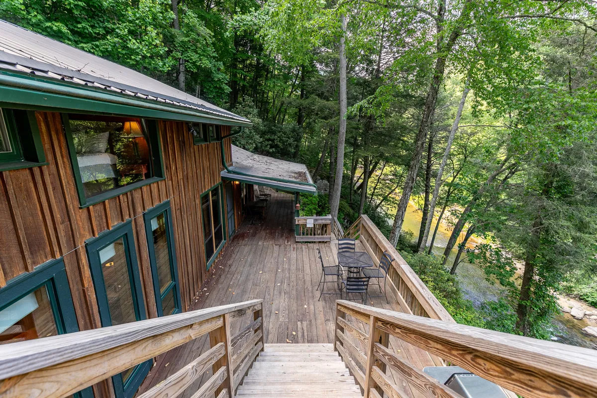 A serene wooden deck overlooks the flowing Watauga River, surrounded by lush green foliage. Comfortable outdoor seating invites guests to sit and enjoy the tranquil atmosphere, with the river’s edge just beyond the railing. The early morning light and mist create a peaceful, immersive connection to nature.
