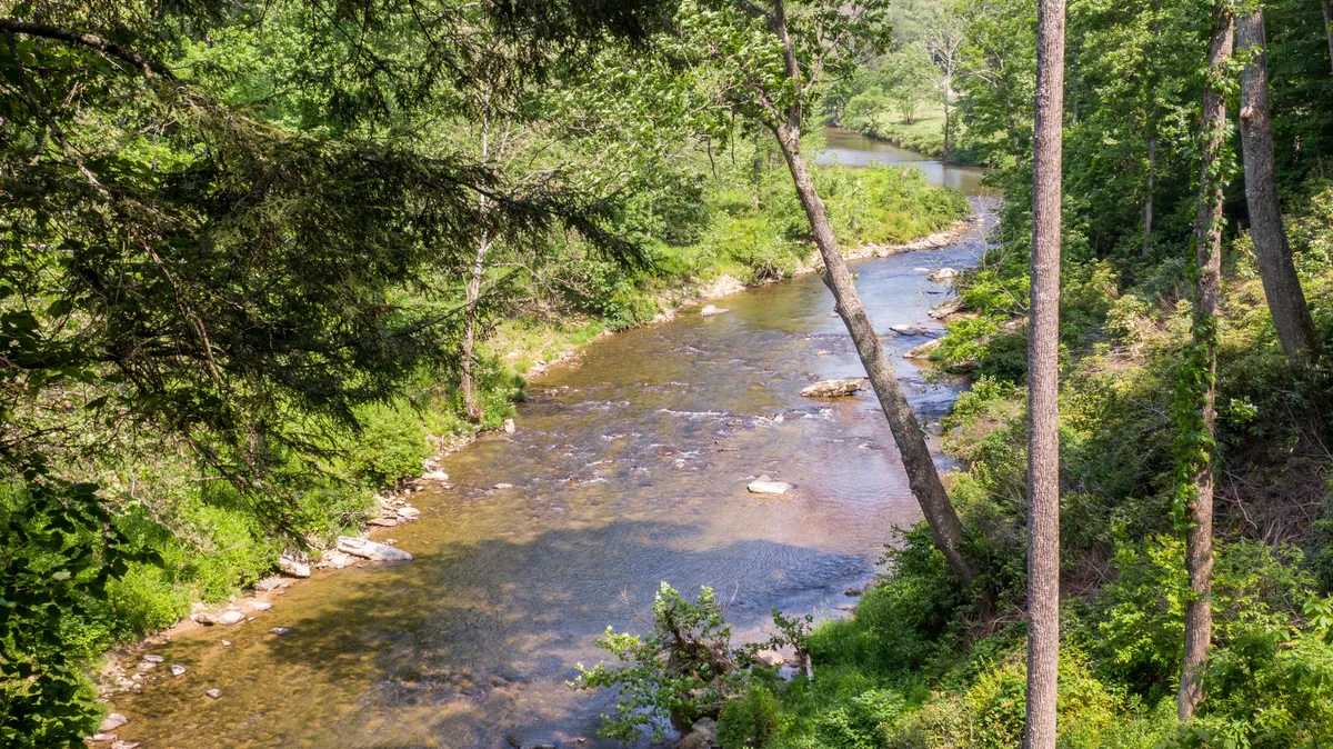 A serene riverside view featuring a flowing river, trees, and a rocky riverbed. The natural landscape invites outdoor activities like riverside chats, fishing, or simply relaxing by the water.