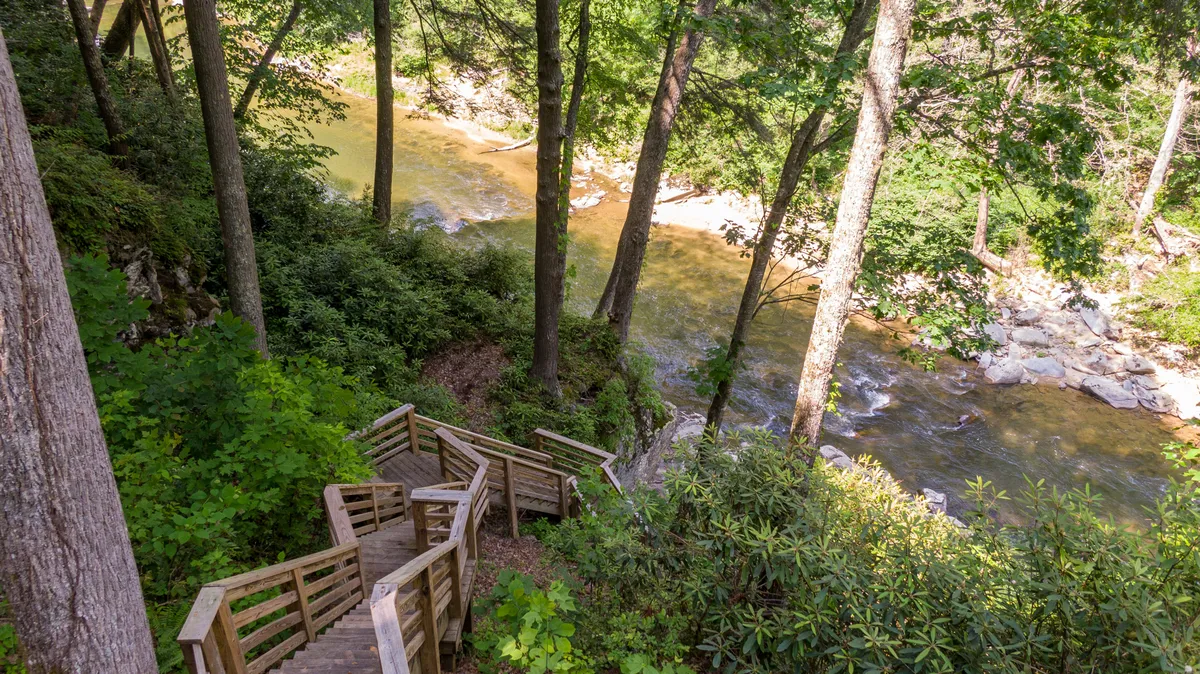 A wooden staircase descends through dense greenery toward the rushing river below. Tall trees and the flowing water create a sense of privacy and immersion in nature, while the stairs provide obvious access directly to the river’s edge.