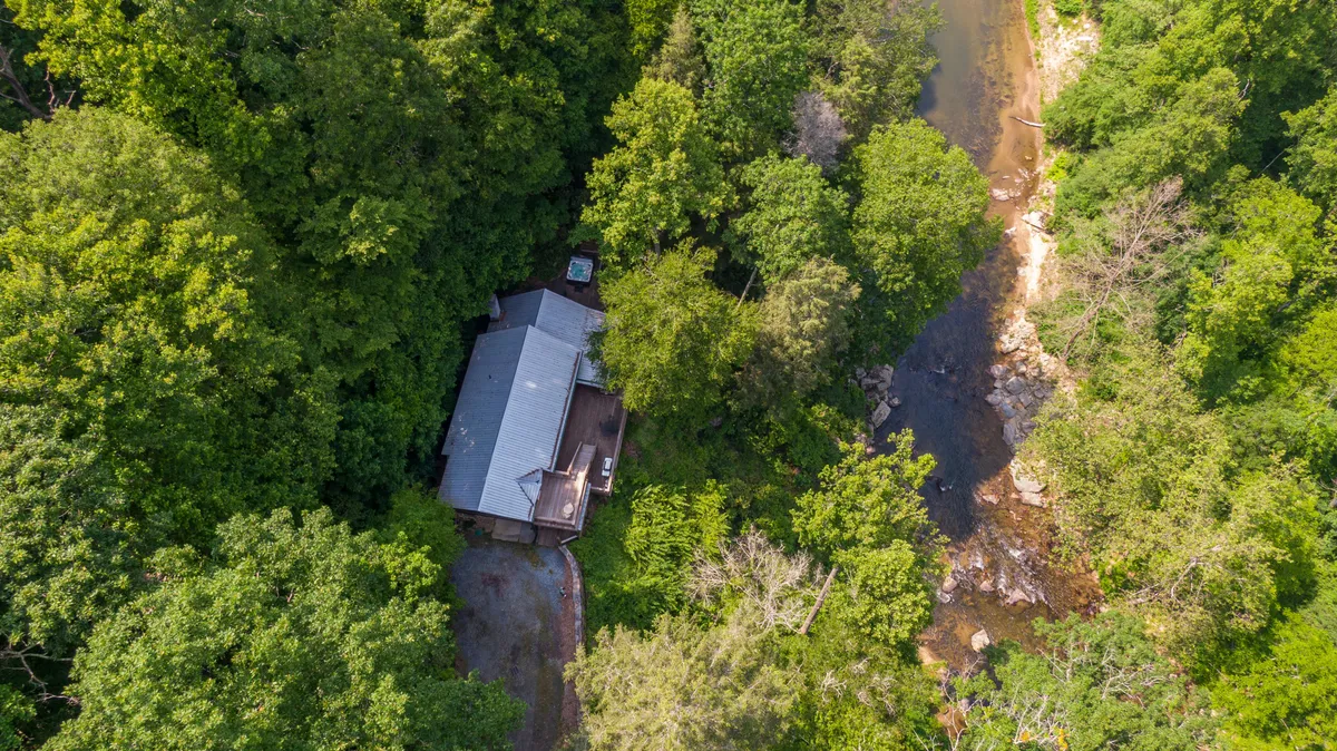 The rustic house is nestled among tall trees beside a peaceful creek, highlighting both the home's proximity to water and its secluded, wooded surroundings. The area looks ideal for nature walks and outdoor fun.