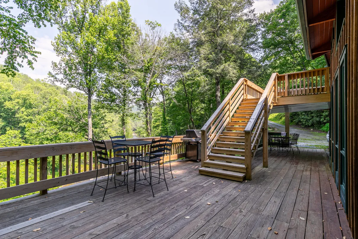 The photo shows a wooden deck with an outdoor seating area, a grill, a staircase, and lush surrounding trees. The space is well-suited for group meals, socializing, and casual gatherings in the fresh mountain air.