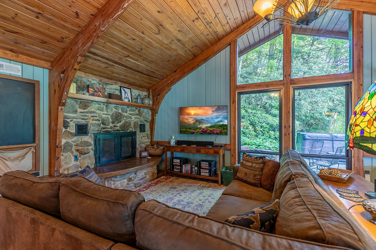 A cozy living room anchored by a large stone fireplace, with wooden beams stretching overhead and large windows overlooking lush greenery. The inviting sectional sofa and a colorful stained glass lamp create a space perfect for relaxing, socializing, or enjoying a movie night. The presence of natural light and views of the outdoors reinforce the riverside setting.