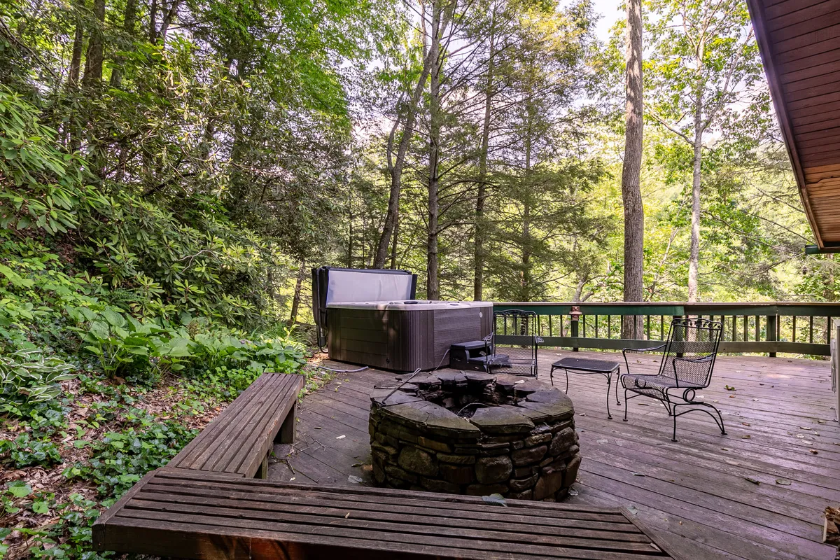 A spacious wooden deck nestled among tall trees, featuring a bubbling hot tub and a stone-ringed fire pit. Comfortable outdoor seating surrounds the fire pit, inviting groups to gather under the open sky. The lush greenery provides a sense of seclusion and immersion in nature.