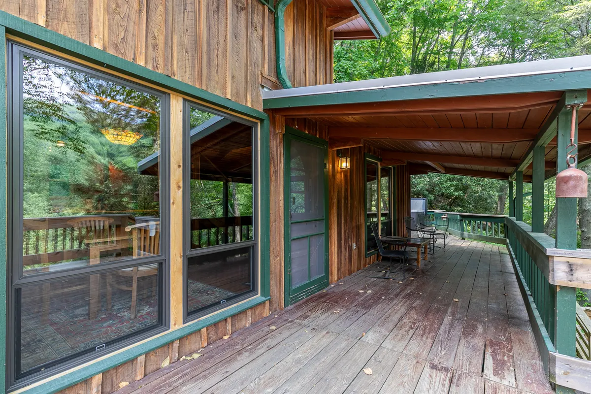 A wooden deck adorned with green trim and outdoor seating, set against a backdrop of tall trees and large windows that connect the indoor and outdoor spaces. The peaceful setting is ideal for enjoying meals, reading, or simply relaxing after a day out.