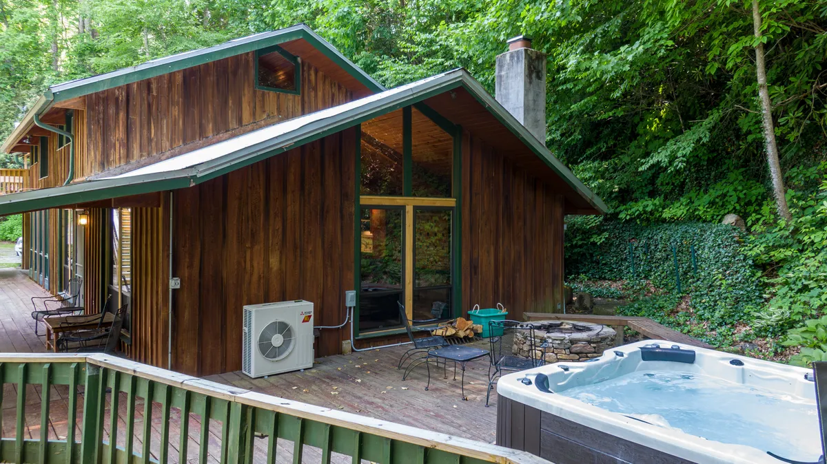 This exterior image showcases a wooden cabin with a welcoming patio, large windows, a hot tub, and outdoor seating. The area is surrounded by trees, evoking a sense of seclusion and natural beauty. The presence of the hot tub and patio seating suggests year-round enjoyment, including cozy winter evenings under the stars or relaxing after a day of adventure.