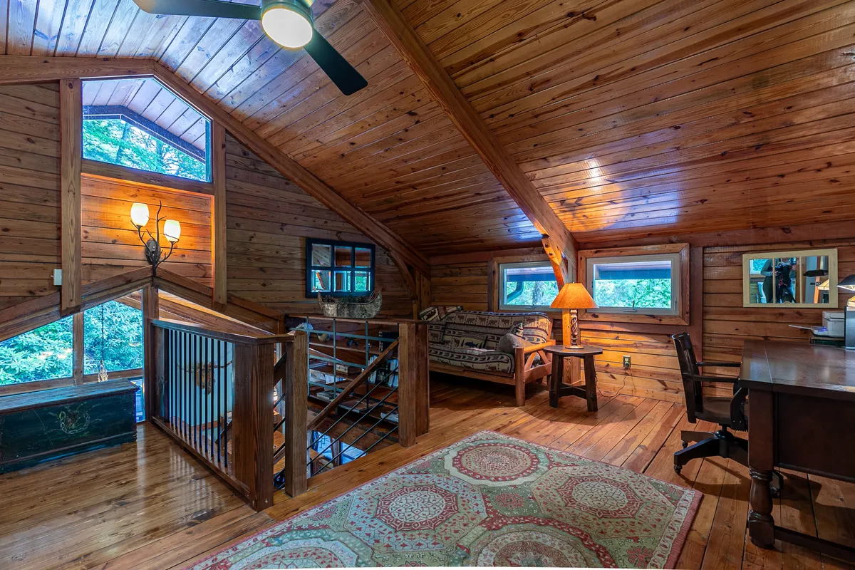 An inviting loft area featuring exposed wooden beams, a comfortable sofa, a work desk with a chair, a decorative lamp, and a cozy area rug. The setting is both functional and relaxing, perfect for work, reading, or creative escapes.