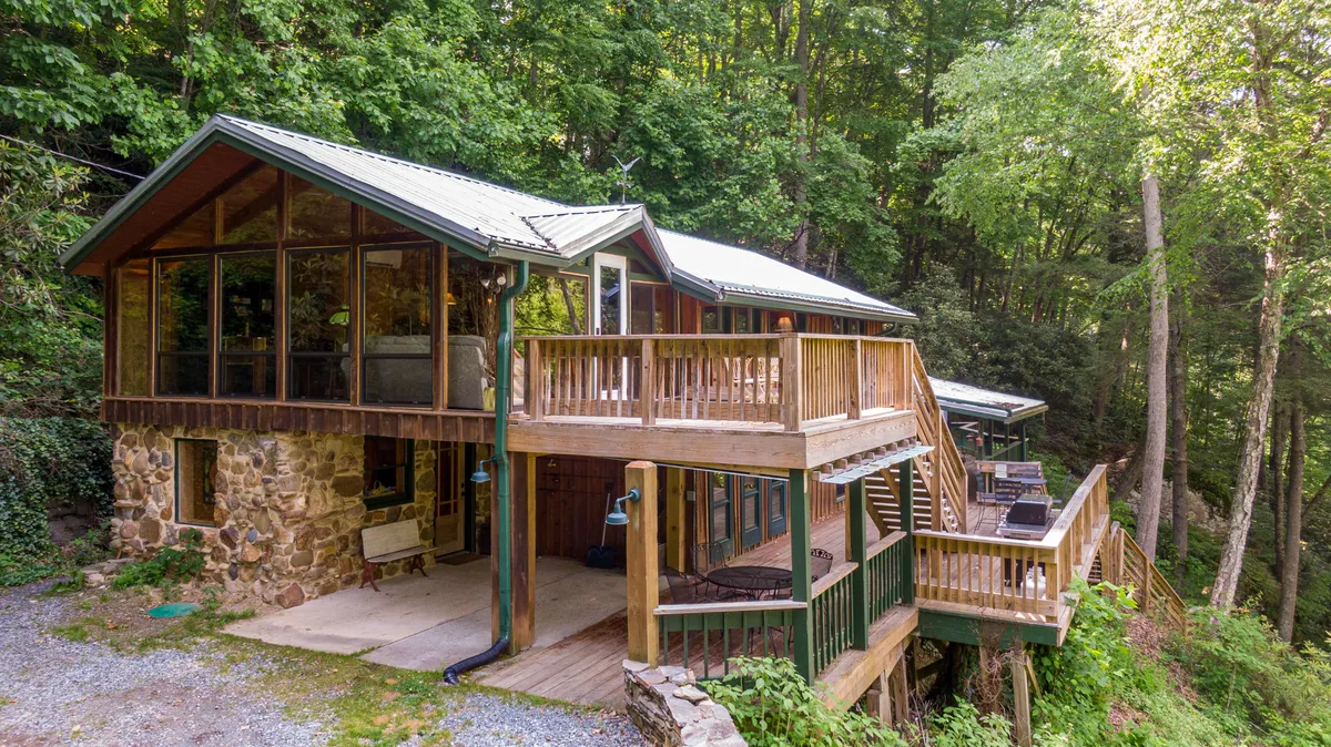 The image showcases the cabin's natural stone facade and expansive glass windows, opening onto a spacious wooden deck nestled among lush greenery. The scene exudes tranquility, with the deck arranged for outdoor dining and relaxation, inviting guests to enjoy sunset views, morning coffee amidst nature, and stargazing under clear mountain skies.