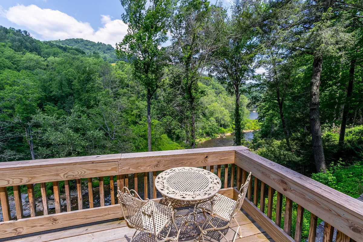 A rustic wooden deck overlooks a lush backdrop of green trees with the river visible in the background. An ornate metal table and chairs suggest an inviting spot for al fresco dining or morning coffee while appreciating the peaceful river view and surrounding wilderness.
