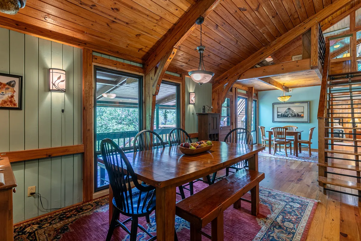 A warm, sunlit dining area with a wooden table ready for group meals, large windows that frame greenery beyond, and rustic wooden beams overhead. The inviting setup is perfect for morning coffees, evening feasts, or board games and conversation.