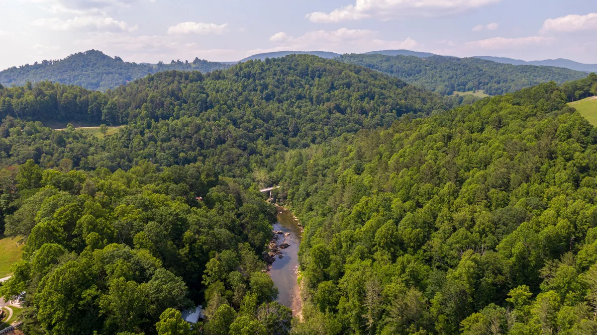 A panoramic view of lush green hills, a meandering river, and distant mountain ranges beneath a wide, open sky. The landscape invites exploration, with the river and forested trails promising hiking, fishing, and picnicking opportunities.
