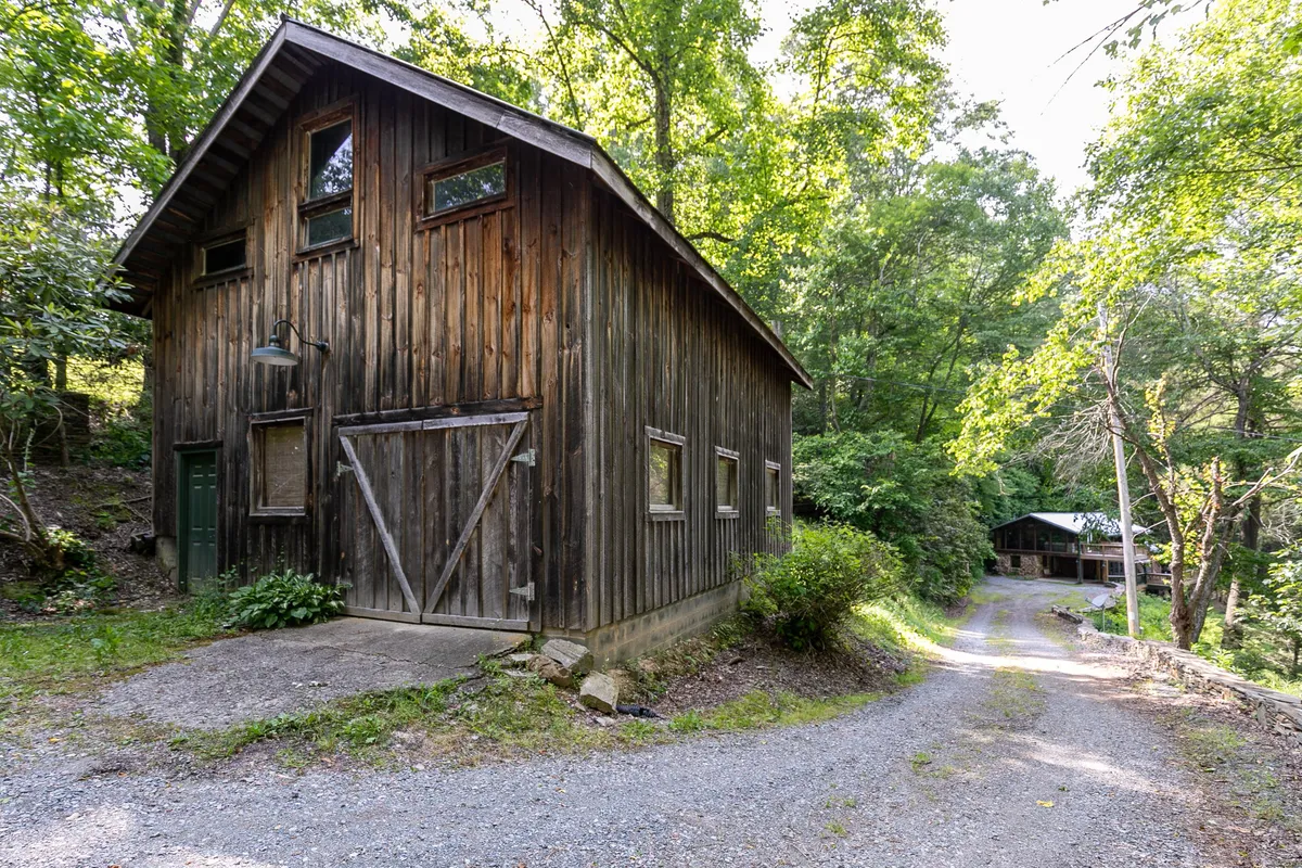 The wooden cabin is nestled among tall trees, with a gravel driveway leading up to it. The setting exudes mountain seclusion and the adventure of arriving at a remote retreat. The scene hints at the journey required to reach such a place, especially in winter conditions.