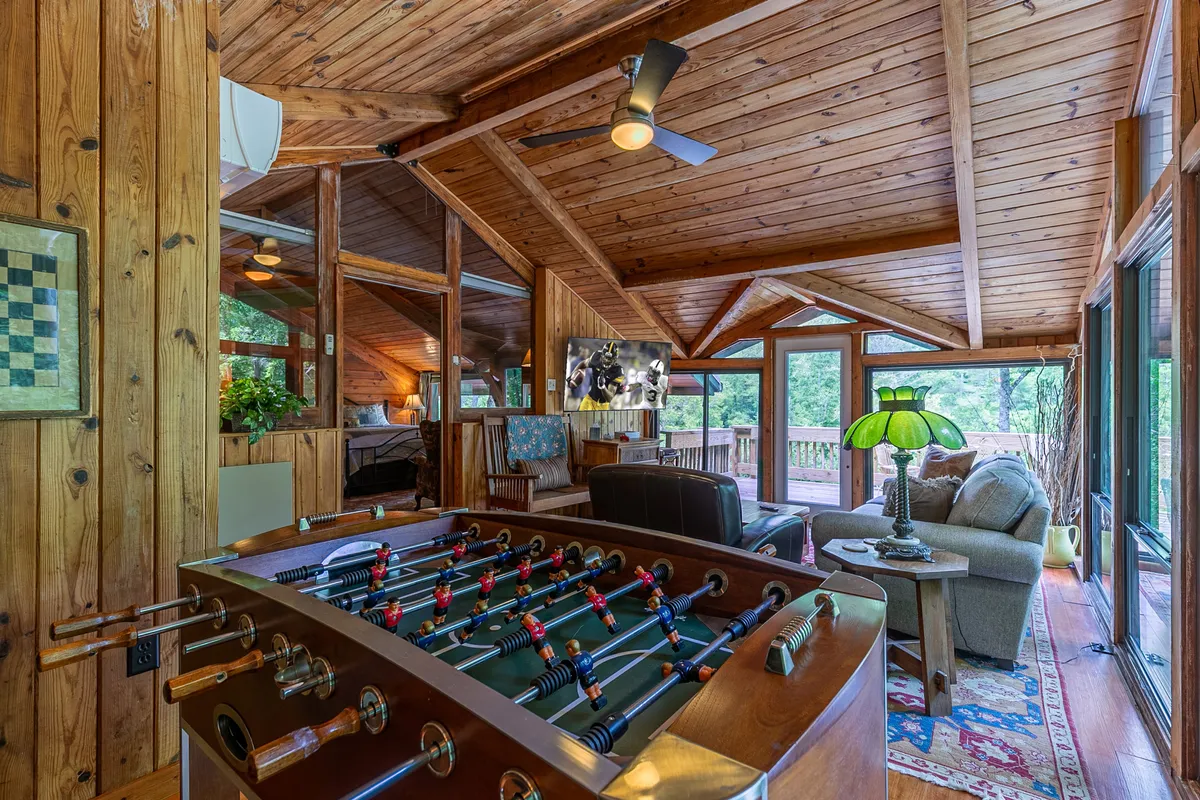 A bright, glass-enclosed game room with a foosball table at the center, comfortable seating arranged near large windows, and a wooden ceiling overhead. The windows frame views of the surrounding greenery, and the space feels casual and inviting, perfect for games and relaxation.